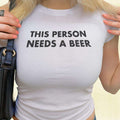 White crop baby tee with bold “THIS PERSON NEEDS A BEER” graphic on front