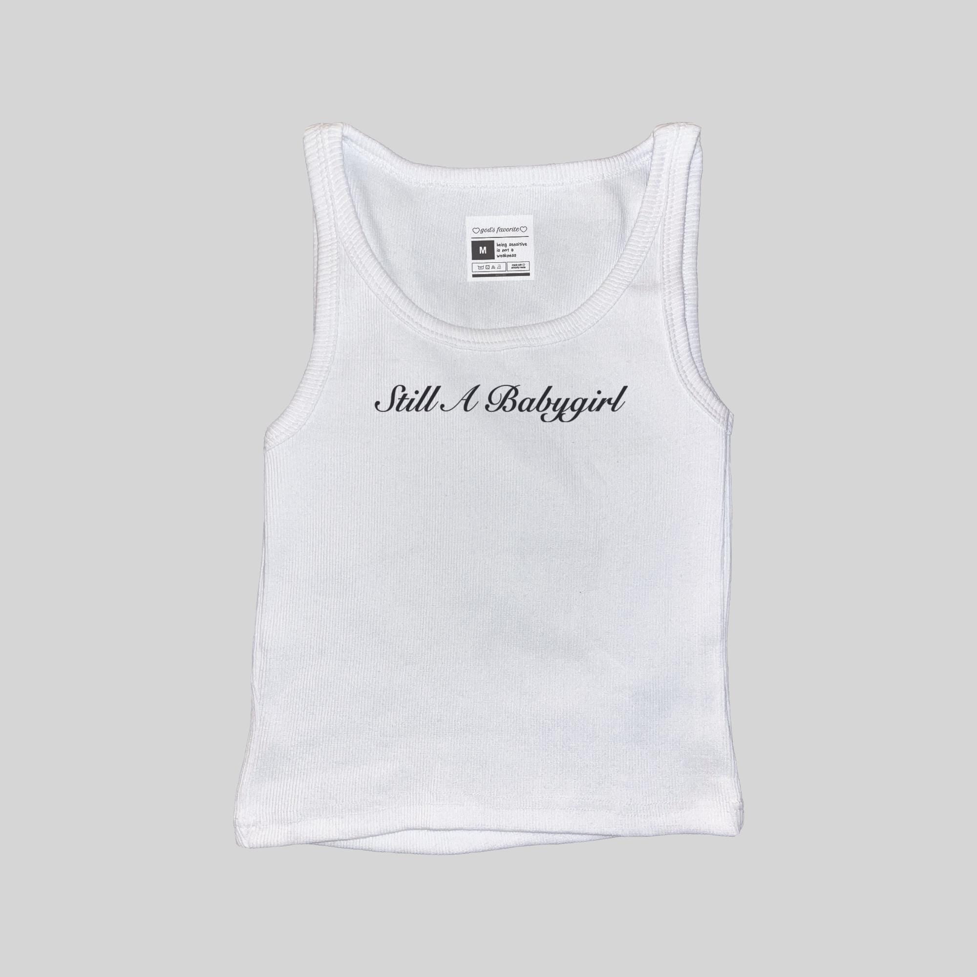 White ribbed sleeveless tank top with front 'Still A Babygirl' script, size tag