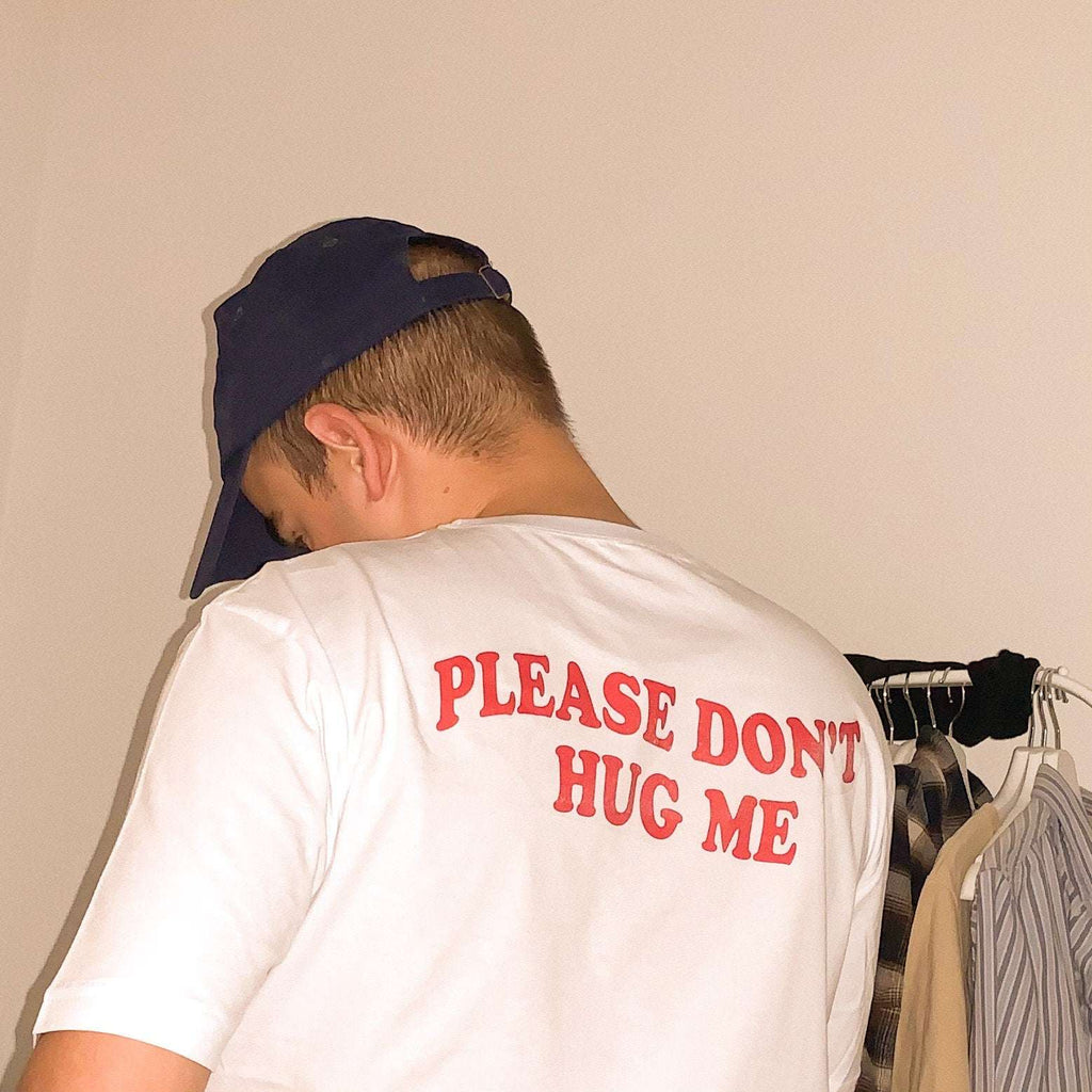 White graphic T-shirt with red “PLEASE DON’T HUG ME” text on the back, worn