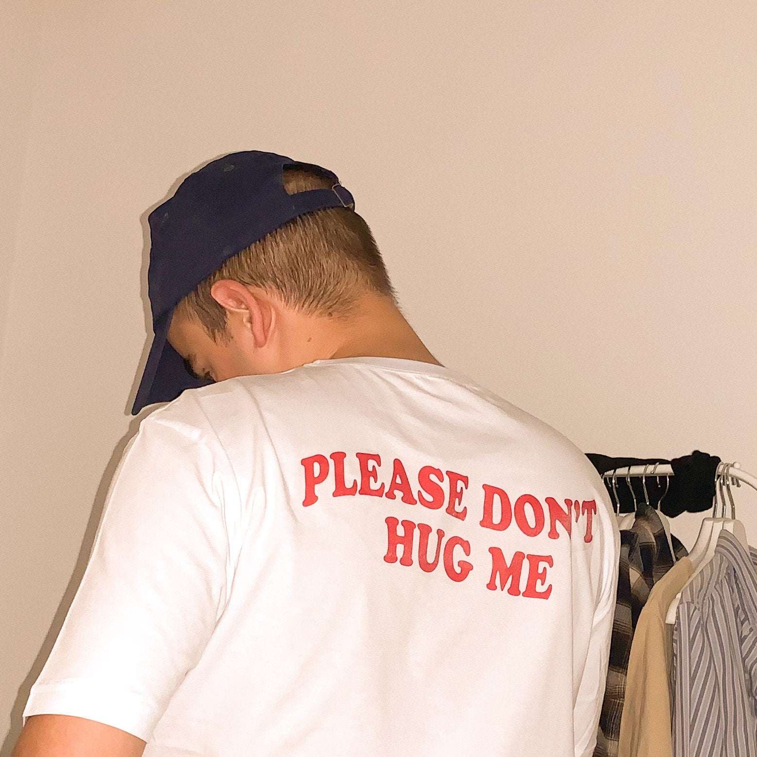 White graphic T-shirt with red “PLEASE DON’T HUG ME” text on the back, worn